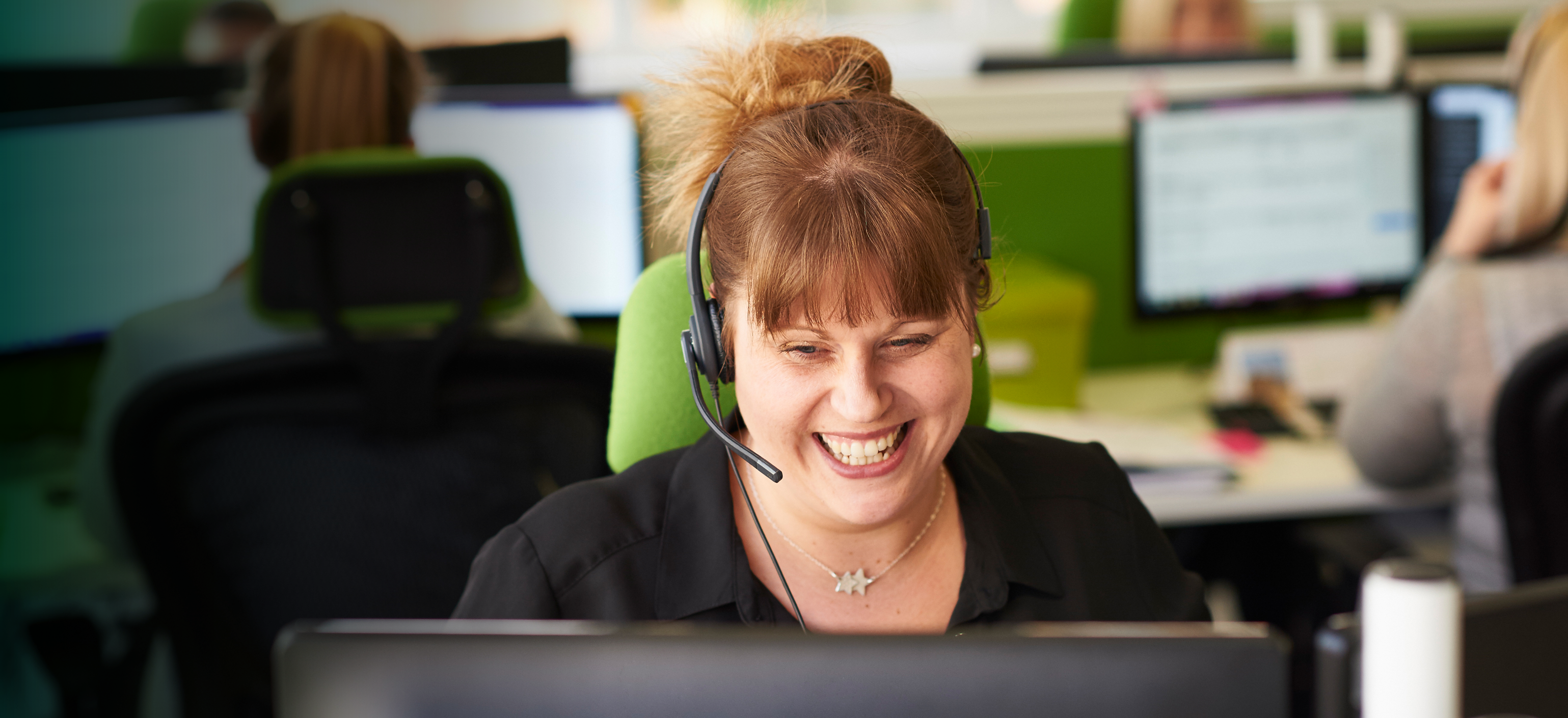 Eine Mitarbeiterin mit einem Headphone sitzt lächelnd vor einem Computer.
