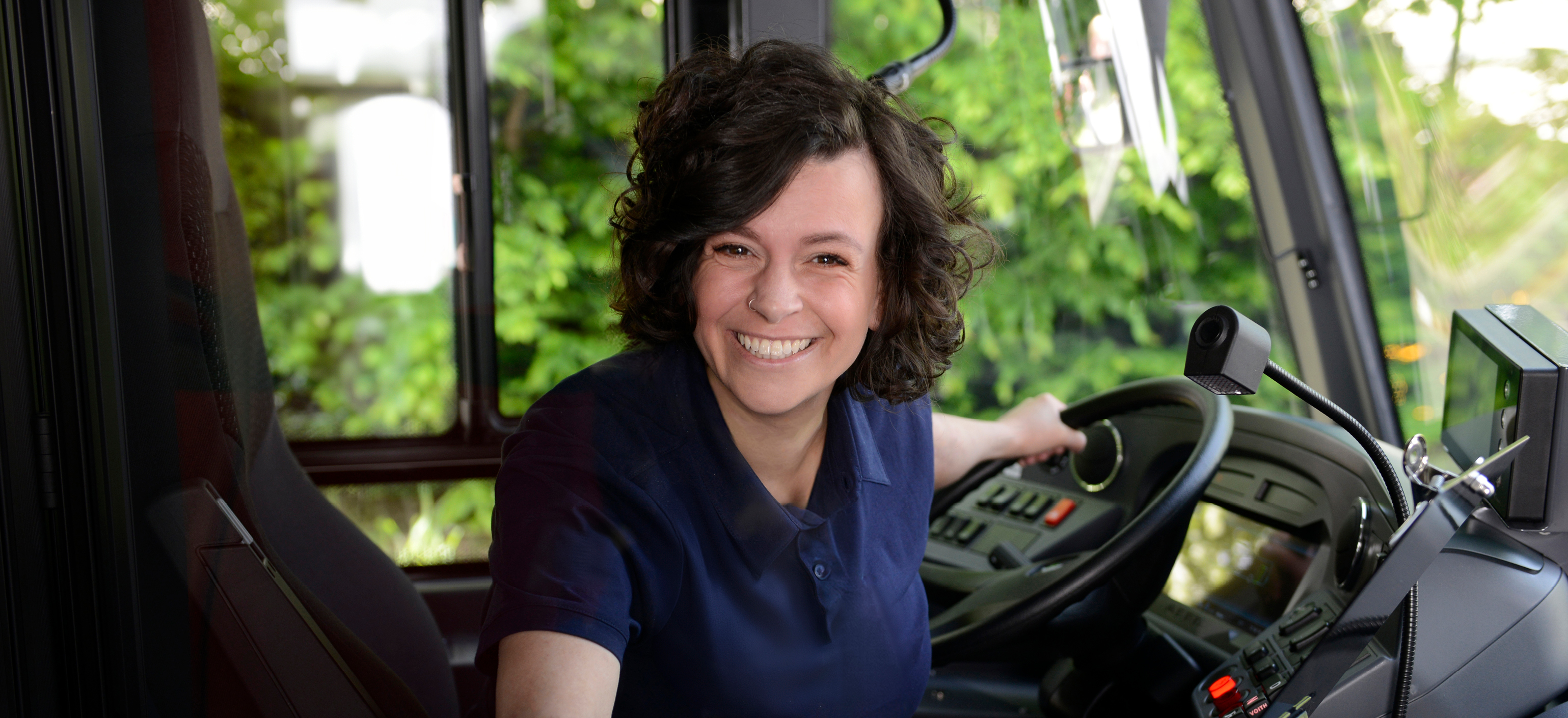 Lächelnde Busfahrerin sitzt am Steuer eines Busses und schaut in die Kamera.